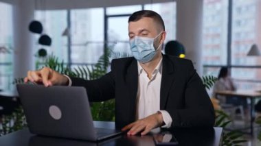 Takım elbiseli ve maskeli bir erkek, dizüstü bilgisayarın önünde oturuyor. Ofiste çalışırken salgın sınırlamalarını takip eden bir adam. Bulanık arkaplan.