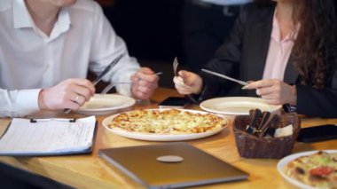 İnsanlar teneffüste kafede yemek yiyorlar. Kadın ve erkek pizza kesiyor. Laptop, telefonlar ve çizelgeler masada..