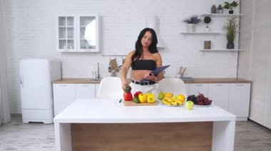 Mutfaktaki çekici kadın sağlıklı bir diyet için ürünler seçiyor. Kadın, önündeki masaya meyve ve sebzeler hakkında notlar alıyor..
