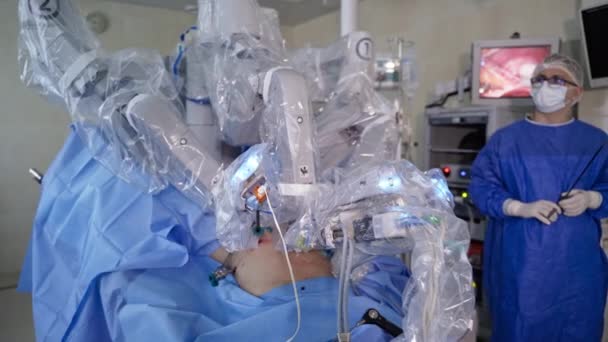 動作テーブルの上に患者を移動するロボットアーム 彼の手のツールを持つ男性外科医は スクリーンの操作に続いています — ストック動画