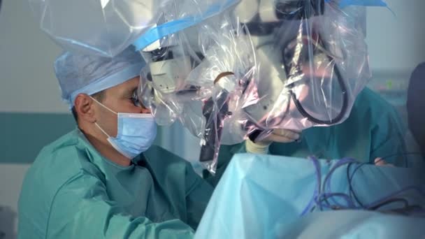 外科医生坐在病人身边 看着显微镜 在手术背景下帮助医生的助手 — 图库视频影像