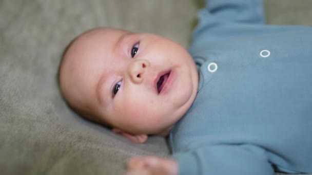 ベッドの上に寝ている大人っぽい赤ん坊 青いシャツを着た赤ん坊のスウィートウィングが近づいています グレーブランケットの背景 — ストック動画
