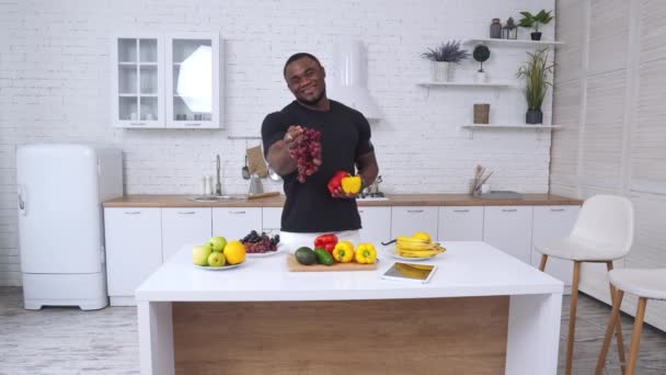 キッチンにオーガニックフード付きの強いフィット感のアフロ系アメリカ人 カメラにブドウを提案し 次に唐辛子を提案する筋肉男 健康的なダイエットコンセプト — ストック動画