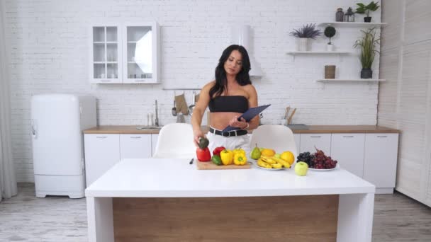 キッチンの魅力的な女性は 健康的な食事のために製品を選択します 女性は彼女の前にテーブルの上の果物や野菜についてのメモを取る — ストック動画