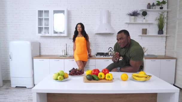 大肌肉的成年人坐在桌旁吃葡萄 穿着橙色迷你裙的女士站在后面 男人向女人招手 她走到餐桌前 端着水果和蔬菜 — 图库视频影像