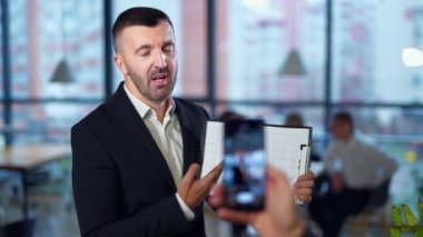 Orta yaşlı işveren kameraya konuşur ve çizelgeyi gösterir. El ele tutuşan telefon ön planda bulanık görüntü alıyor.
