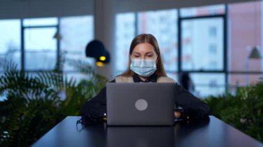 Bilgisayarına bir şey yazan maske takan kadın bir ofis çalışanı. Bayan daha iyi görebilmek için ekrana yaklaşıyor. Ofis arkaplanı bulanık.