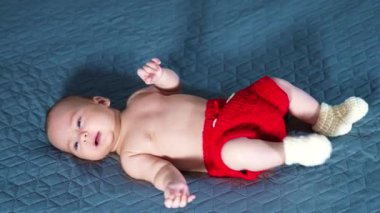 Kırmızı şortlu ve beyaz çoraplı aktif bir çocuk. Beyaz bebek yatakta yatarken ayaklarını neşeyle hareket ettiriyor. Gri arkaplan.