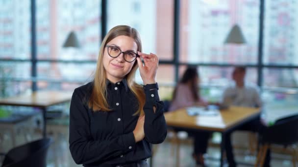 自信而成功的年轻女士戴着眼镜笑着对着相机 站在办公室里的妇女戴上眼镜 后台办公室里的人 — 图库视频影像