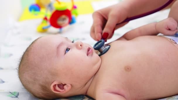 儿科医生的手把听诊器放在孩子的胸口上 婴儿看上去很有兴趣 对医生笑了 模糊的背景 — 图库视频影像