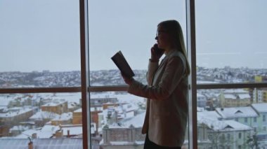 Uzun saçlı bir kadın telefonda konuşuyor ve önünde bir defter tutuyor. Arkaplanda kış manzaralı panoramik pencere.