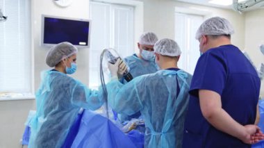 Modern klinikte ameliyat yapan profesyonellerden oluşan bir ekip. İki doktor hastanın üzerinde ağır bir tıbbi cihaz taşıyor. Önplanda erkek doktor süreci izliyor.