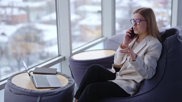 現代のライトオフィスに座っている若い女性起業家 魅力的な女性が電話で話し 微笑んでいる ブラーレッド バックドロップ — ストック動画