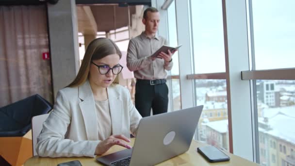 ライトオフィスで働く従業員 デスクにノートパソコンを入力する女性 バックドロップの窓の近くにノートと立っている男性 — ストック動画