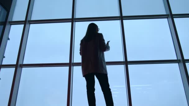 背中に立っている長髪の女性の暗いシルエット パノラマウィンドウの前で電話で話す女性 ローアングルビュー — ストック動画