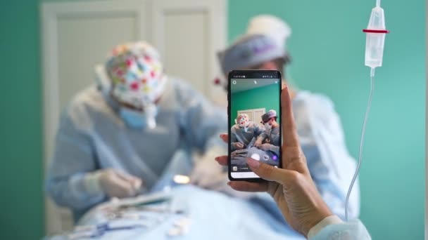 スマートフォンを持って手術のビデオを撮る女性の手 2人の外科医がバックドロップで爆発作動 — ストック動画