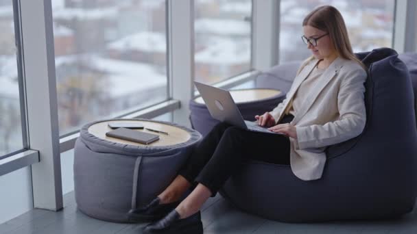 ファッショナブルなオフィスの前提で快適な職場 ラップトップで働くビーンバッグの椅子に座っている女性 パノラマウィンドウの背景 — ストック動画