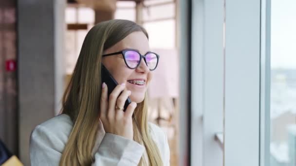 微笑的女士在电话里说话 关闭一个积极的年轻女士的肖像站在大的全景窗口附近 — 图库视频影像