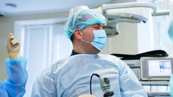 デバイス付きの男性外科医は 患者の上に立っています 薄いワイヤーを持っている女性アシスタントは 医師の隣にあります バックドロップ時の医療機器 — ストック動画