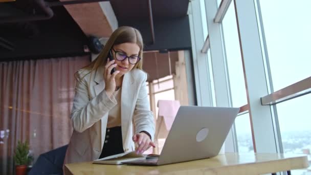 ラップトップ付きの机の上に立っている美しい働く女性 忙しい女性が電話で話し メモをチェックします オフィスの背景 — ストック動画