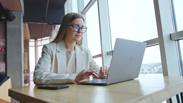 若い女性は机の上に座っているラップトップで働いている メガネの女性は 意図的に画面を見ています パノラマウィンドウの職場 — ストック動画