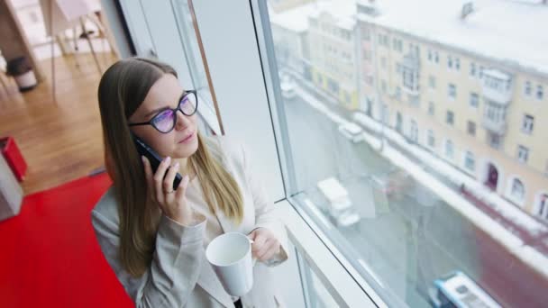 お茶のカップで電話でビジネス話をする スマートフォンでチャットウィンドウのそばに立っている若い女性 高角度ビュー — ストック動画