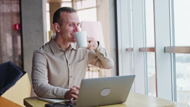 レジリエントな若者はノートパソコンを持って机に座り お茶を飲んでいる 職場で昼食休憩をしている男性 オフィスバックドロップ — ストック動画