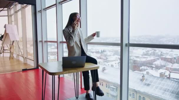 テーブルに座って お茶を飲んで電話で話している幸せな女性 オフィスでランチ休憩をしているビジネスレディ バックドロップのシティスケープ — ストック動画