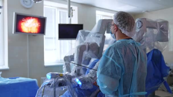 オペレーティングエリアをスクリーンで見ている男性外科医 壁のモニターは手術中のロボットアームの仕事を示しています — ストック動画