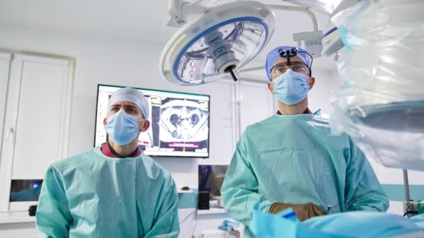 二人の医師が目の前をじっと見つめている チーフ外科医は患者の体に金属製の道具を挿入する ローアングルビュー — ストック動画