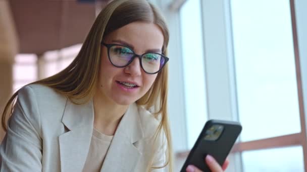 戴着眼镜和手镯的年轻女子手里拿着电话 看着笔记本电脑 一个微笑的女士在办公室工作的画像 靠近点 — 图库视频影像