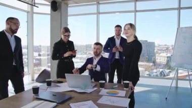 İşletme şefi dizüstü bilgisayarın önünde oturuyor. Ofis ekibi liderlerinin etrafında toplanıyor. Arkaplanda şehir manzaralı panoramik pencereler.
