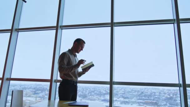 英俊而苗条的男人站在窗前看他的笔记本 办公室工作人员做一些笔记 背景下的城市景观 — 图库视频影像