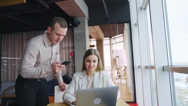 ライトオフィスのジェンダーの同僚は ラップトップを見ています 女性は机に座り 男は彼女の隣に彼の手で携帯電話を持って立っている — ストック動画