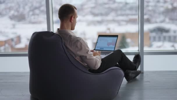 忙碌的男性自由职业者背对着摄像机坐在豆袋椅子上 人类在屏幕上有图表的笔记本电脑上工作 背景上城市景观模糊的窗户 — 图库视频影像