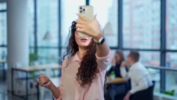 長い暗いカーリーの髪とメガネのチャーミングな女性は 彼女の携帯電話でビデオを取ります バックドロップで働くオフィスチーム — ストック動画