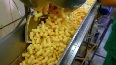 Bir sürü sarı gevrek mısır çubuğu taşıma hattına düşer. Fabrika işçisi aperatifleri kemer boyunca itiyor. Gıda fabrikası üretim konsepti.