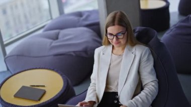 Gözlüklü hoş görünümlü kadın arkasına yaslanıyor ve bacaklarında dizüstü bilgisayarla rahat bir şekilde koltuğa oturuyor. Modern ofis işi konsepti.