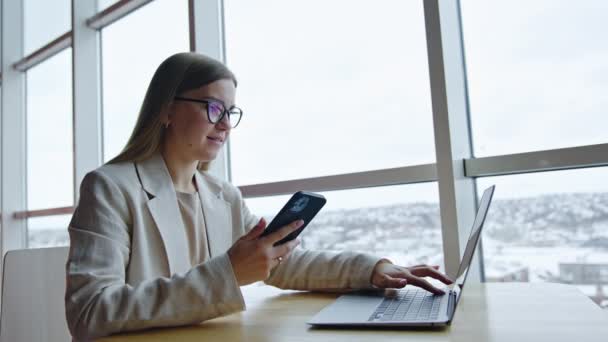 年轻妇女坐在靠窗的办公桌前 用小玩意 女职员用智能手机进行商务交流 — 图库视频影像