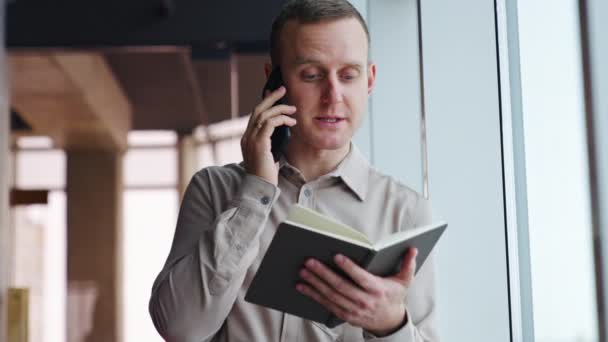 灰色のシャツを着た男が電話をする 男性の従業員は彼の前に紙のノートを保持し 電話で話す — ストック動画