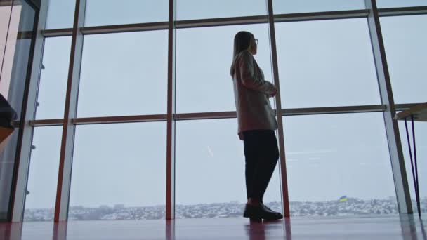 スーツの長髪の女性は パノラマの窓の近くを歩いています 若い女性が街のパノラマを見ている ローアングルビュー — ストック動画