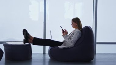 Ceketli ve kot pantolonlu bir kadın laptopuyla işten uzaklaştırılmış. Genç bir kadın telefonunda bir şey kaydırıyor. Panoramik pencere arkaplanı.