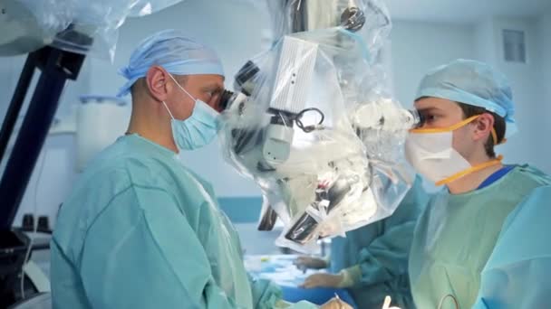 外科医のカップルは 双眼鏡を調べる現代機器の両側に立っています 金属製の器具を持つ医師の手は患者の体を扱う装置の下で動きます — ストック動画