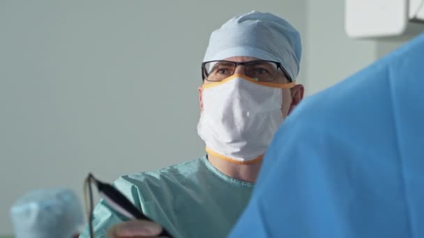 大人の男性外科医はスクリーン上にまっすぐ見えます 眼鏡の医師は 患者の体に入力したデバイスを保持します — ストック動画