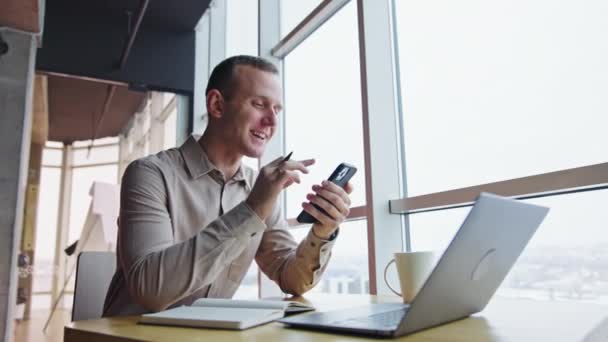 Mann Sitzt Schreibtisch Schaut Auf Sein Telefon Spricht Und Lächelt — Stockvideo