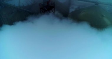 Tahıl asansöründeki borulardan yoğun beyaz duman geliyor. Gri sisli bir günde metal tankların hava görüntüsü..