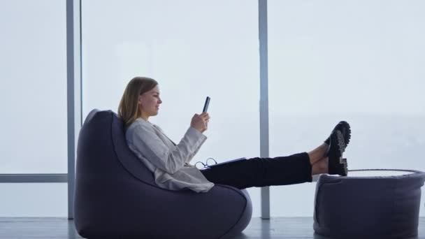 若い女性はオットマンに彼女の足を伸ばすビーンバッグチェアに快適に座っています 女性は携帯電話と笑顔を持っています ラップトップとメガネが彼女のラップの上にあります 背景にあるパノラマウィンドウ — ストック動画