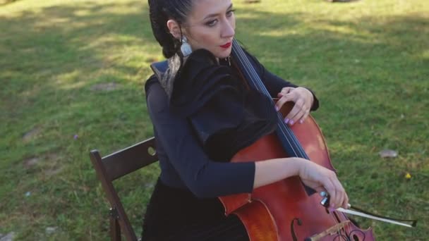女士穿着黑色连衣裙 长长的耳环和红色口红在户外弹奏大提琴 女音乐家表演音乐并享受这一过程 自然背景 — 图库视频影像