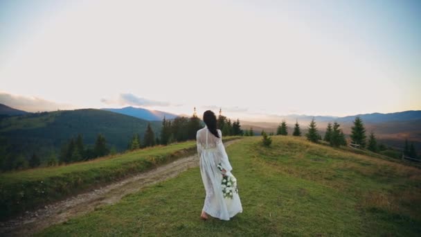 穿着透明白色衣服的赤脚女士在山上散步 长发女人手里拿着白花 以冷杉为背景的高山风景 — 图库视频影像