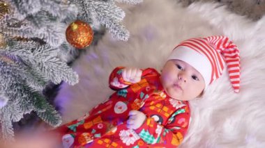 Kırmızı beyaz şapkalı, ciddi bir erkek bebek bembeyaz ekose kumaşın üzerinde yatıyor. Süslü Noel ağacının yanındaki komik çocuk. Yukarıdan görüntüle.
